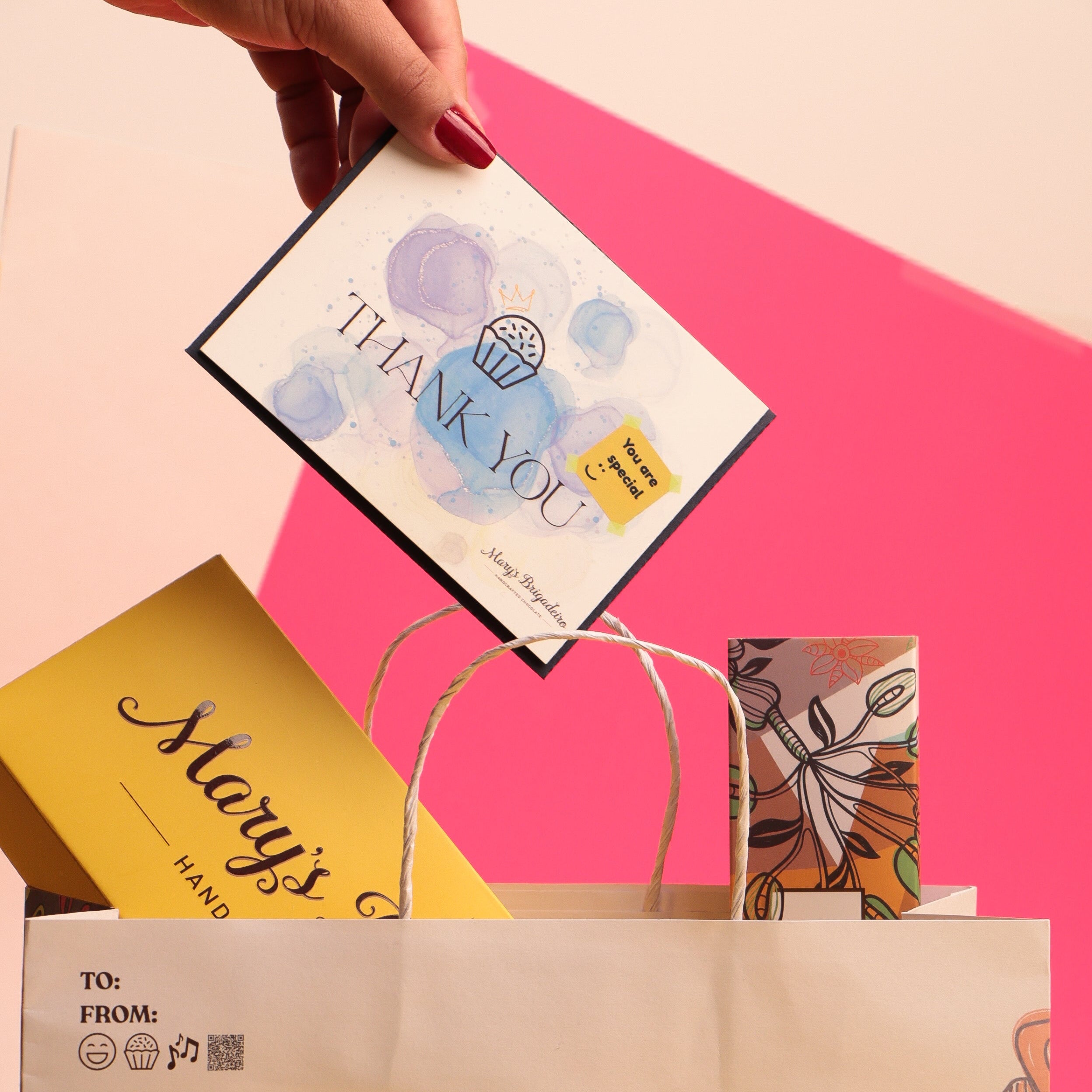 Hand holding a 'Thank You' card over a Mary's Brigadeiro gift bag with colorful packaging.