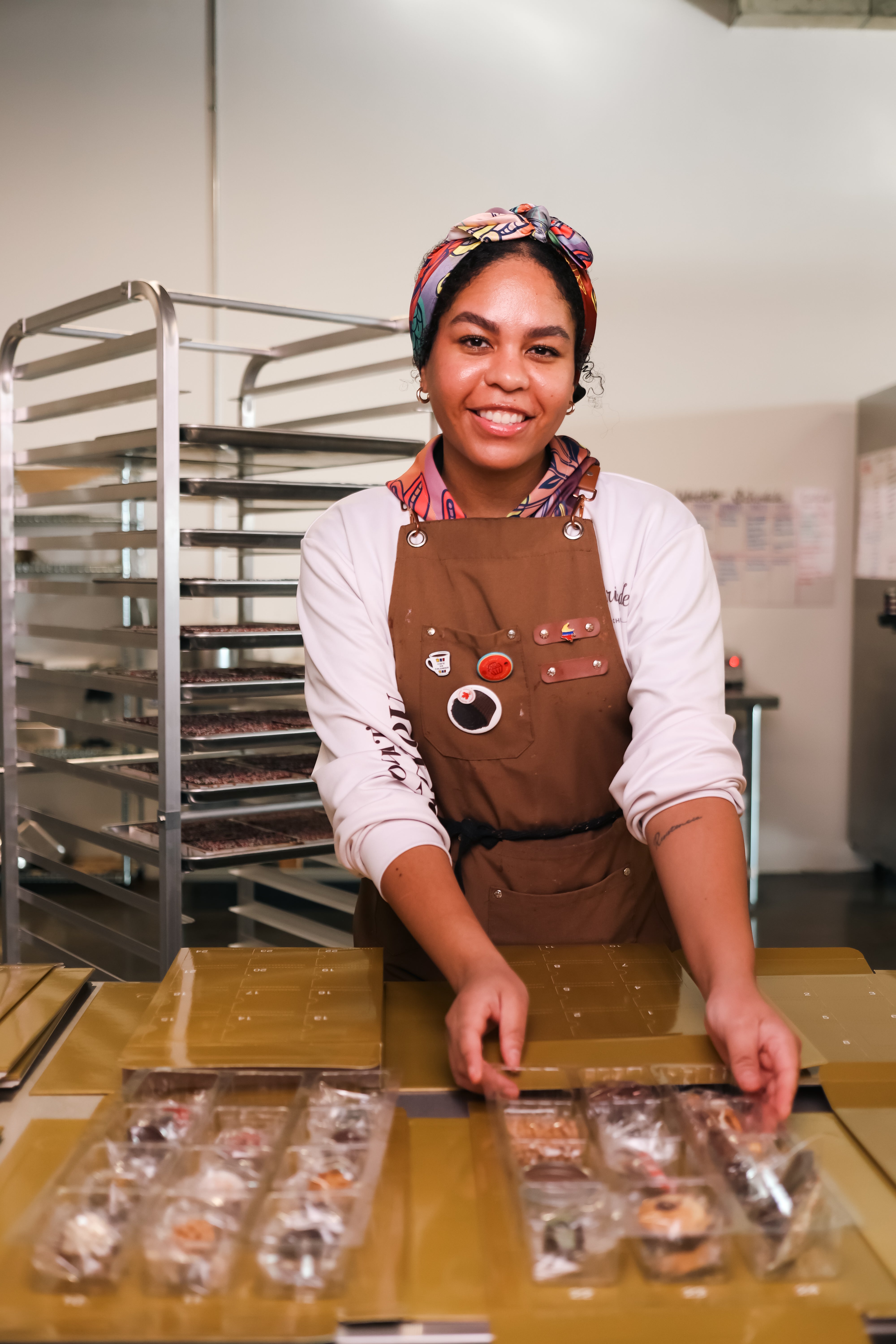 Person wearing a brown apron and headscarf, standing behind a counter with food packages in a kitchen setting.