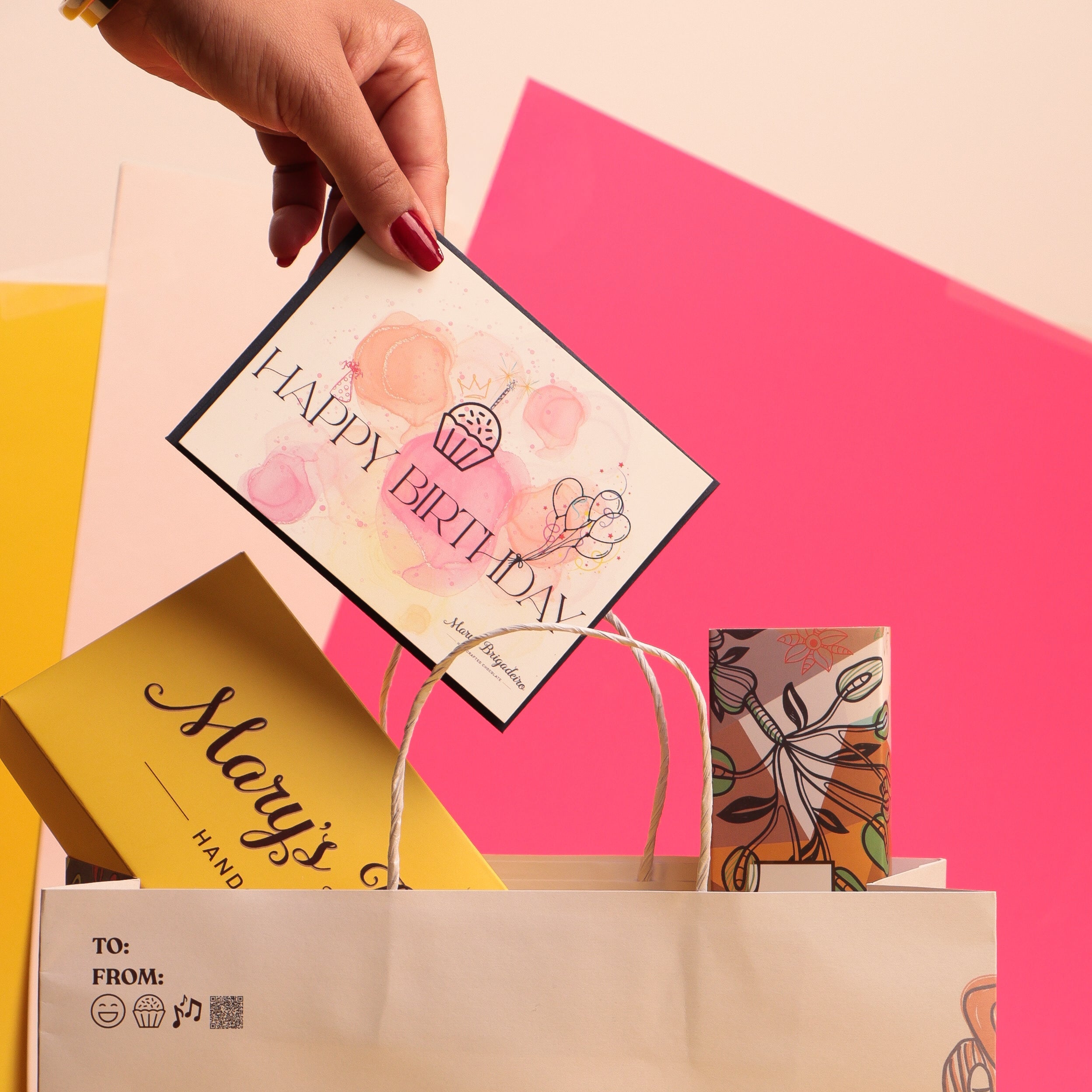 Hand holding a 'Happy Birthday' card over a Mary's Brigadeiro chocolate bag with colorful gift bags in the background.